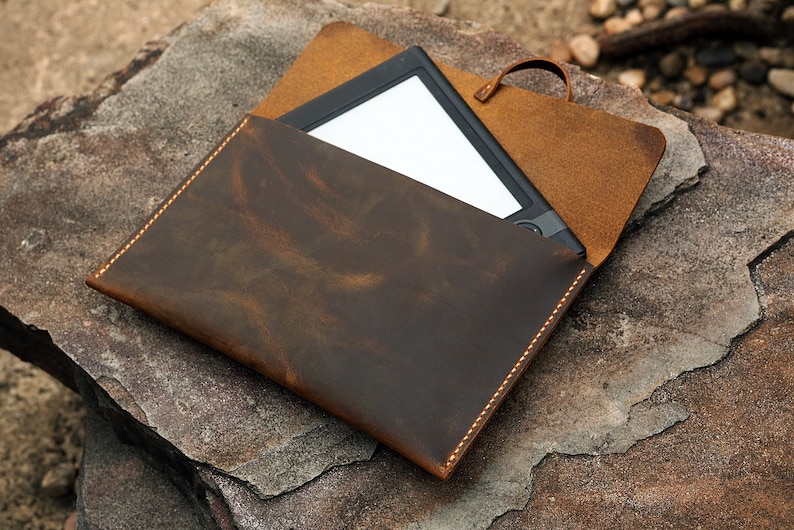 May include: A brown leather case with a flap and a stitched edge. The case is open and a black e-reader is inside.