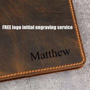 Op de afbeelding: Een donkerbruine lederen portemonnee met zichtbare stiksels langs de randen. Het oppervlak toont de gegraveerde naam Matthew en de gedrukte tekst FREE logo initial engraving service in een wit schreefloos lettertype op het leer.