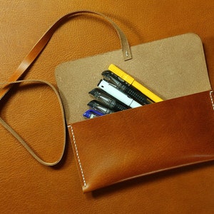 May include: A brown leather pencil case with a strap. The case is open and contains several pens and pencils, including a yellow highlighter.