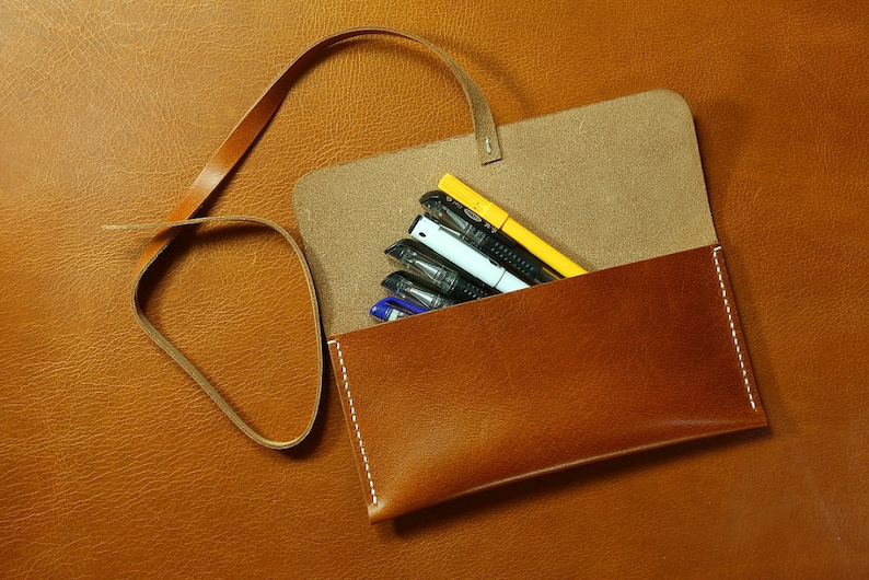May include: A brown leather pencil case with a strap. The case is open and contains several pens and pencils, including a yellow highlighter.
