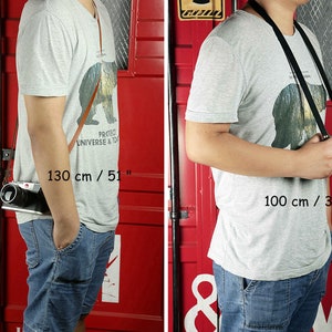 May include: Two people wearing gray t-shirts with a bear graphic and blue jeans are shown wearing camera straps. The strap on the left is brown leather and measures 51 inches long. The strap on the right is black and measures 39 inches long.  "Protect Universe & Today" is printed on the t-shirt.