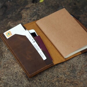 May include: A brown leather notebook cover with a built-in pocket for a phone or passport. The cover has a flap that closes with a snap. The notebook is open and has a blank page visible.