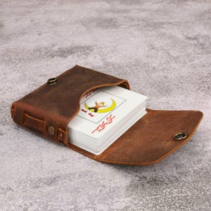 May include: A dark brown leather case with a snap button closure containing a deck of playing cards. The top card features a colourful illustration of a clown sitting on a yellow crescent moon with red text printed vertically along the side.