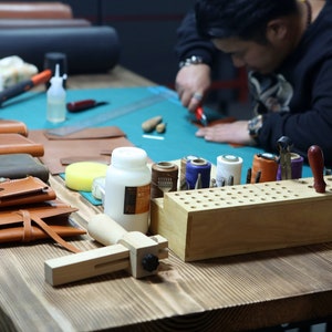 May include: A leather craftsman works at a table with various tools and supplies, including leather pieces, thread, and a wooden tool holder. There are several finished leather goods, such as wallets and pouches, on the table.