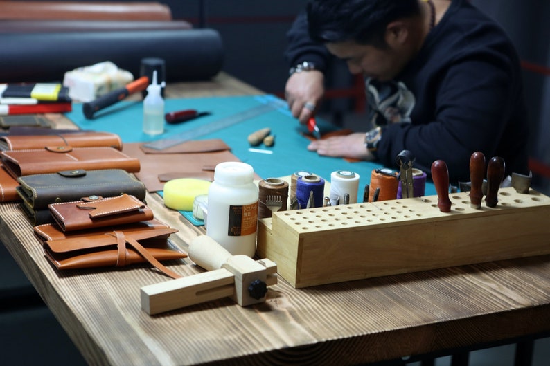 May include: A leather craftsman works at a table with various tools and supplies, including leather pieces, thread, and a wooden tool holder. There are several finished leather goods, such as wallets and pouches, on the table.