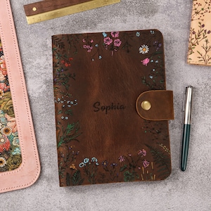 May include: A dark brown leather journal featuring a floral print with pink, blue, and white flowers. The name Sophia is engraved in the center. A leather strap with a brass snap closure secures the cover. A silver pen is placed to the right.