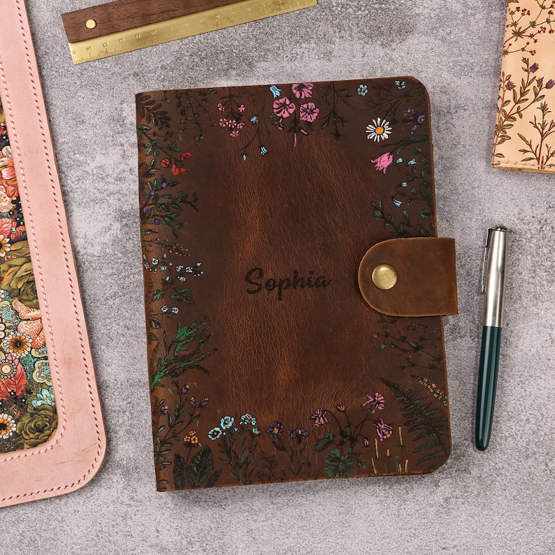 May include: A dark brown leather journal featuring a floral print with pink, blue, and white flowers. The name Sophia is engraved in the center. A leather strap with a brass snap closure secures the cover. A silver pen is placed to the right.