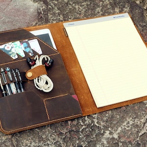 May include: A brown leather portfolio with a notepad, pen holders, and a pocket for a tablet or phone. The portfolio has a flap that closes with a button.