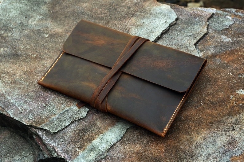 May include: A brown leather envelope with a flap closure and a leather strap. The envelope is tied with a leather strap and has a distressed look.