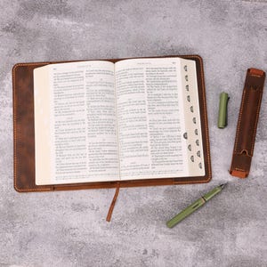 May include: An open book with a brown leather cover and a matching bookmark. A green fountain pen, a pen holder, and a pen cap are next to the book. The book is open to Psalm 119.