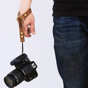 May include: A brown leather camera wrist strap with a brass buckle and two brass rivets. The strap is attached to a black DSLR camera with a lens.