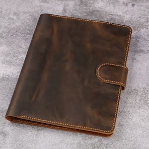 May include: A brown leather binder with a flap closure and a stitched edge. The binder has a distressed look and is made of high-quality leather.