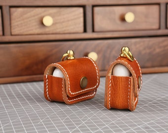 May include: Two small brown leather cases with white stitching and brass hardware. Each case has a small white object inside and a brass loop at the top.
