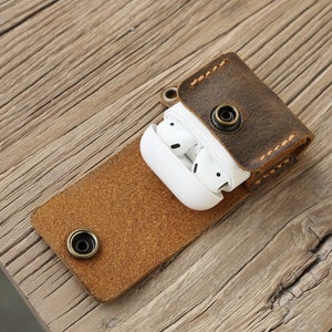 May include: A brown leather AirPod case with a snap closure, holding white wireless earbuds. The case is open, revealing the earbuds. The case is on a wooden surface.