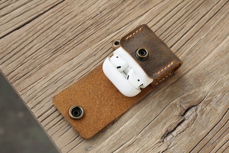 May include: A brown leather AirPod case with a snap closure, holding white wireless earbuds. The case is open, revealing the earbuds. The case is on a wooden surface.