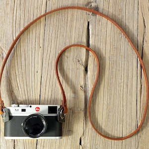 May include: A brown leather camera strap with a silver camera attached. The camera is a Leica M with a black body and a silver lens.