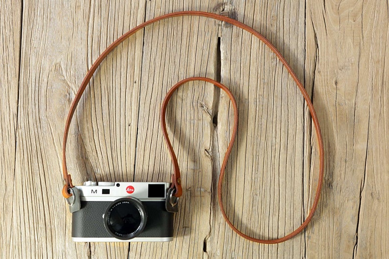 May include: A brown leather camera strap with a silver camera attached. The camera is a Leica M with a black body and a silver lens.