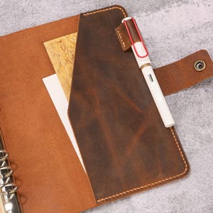 May include: A brown leather binder featuring a metal ring mechanism, a side pocket, and a snap button closure. A white fountain pen with a red clip is inserted into a leather pen loop attached to the cover, holding a sheet of paper inside.