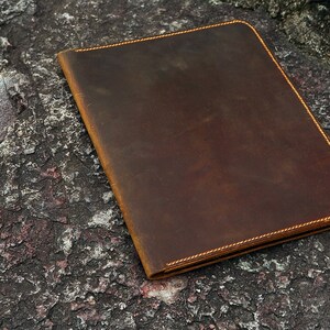 May include: A brown leather folder with a stitched edge. The folder is lying on a rough, textured surface.