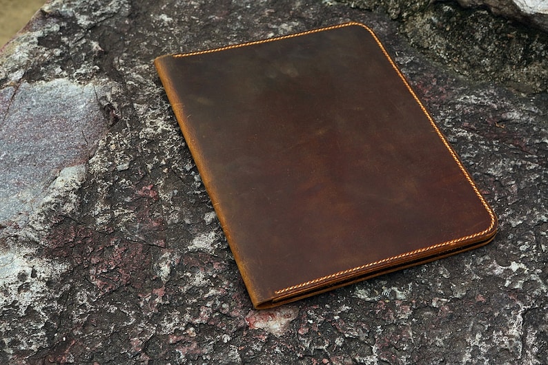 May include: A brown leather folder with a stitched edge. The folder is lying on a rough, textured surface.