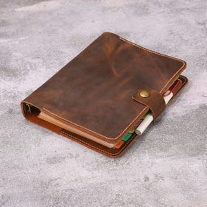 May include: A brown leather binder with a snap button closure and visible stitching along the edges. The binder contains multi-colored index tabs and a white pen tucked into the side loop, showcasing a textured surface and a metal ring mechanism.