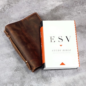 Op de afbeelding: Een witte ESV Study Bible met oranje randen en een bruine leren hoes. De hoes is open en toont de titel van het boek en de woorden "English Standard Version". De leren hoes heeft een gespsluiting.