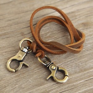 May include: A brown leather lanyard with two antique brass-colored swivel clasps. The leather strap is looped and knotted, and the clasps are attached to the ends. The lanyard is designed to hold keys or other small items.