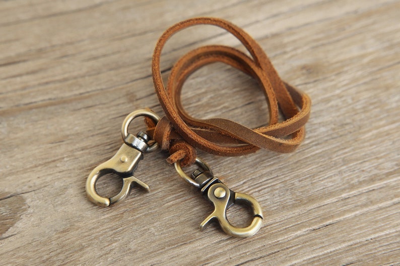 May include: A brown leather lanyard with two antique brass-colored swivel clasps. The leather strap is looped and knotted, and the clasps are attached to the ends. The lanyard is designed to hold keys or other small items.