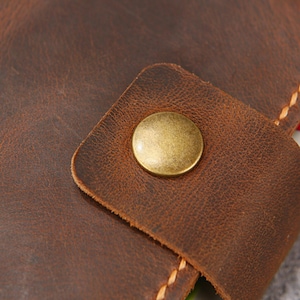 May include: A dark brown leather strap featuring a smooth, circular brass-toned metal snap button fastener. The leather has a visible grain texture and is secured with thick, light-colored stitching along the edges of the strap and base.