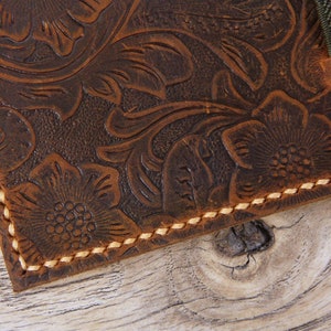 May include: Close-up of a brown leather wallet with a floral embossed design and tan stitching. The wallet has a flap with a green strap.