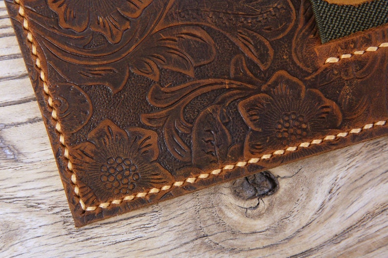 May include: Close-up of a brown leather wallet with a floral embossed design and tan stitching. The wallet has a flap with a green strap.