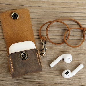 May include: A brown leather AirPods case with a snap closure and a matching leather strap with a metal clasp. Two white AirPods are next to the case on a wooden surface. The case is designed to protect and carry wireless earbuds.