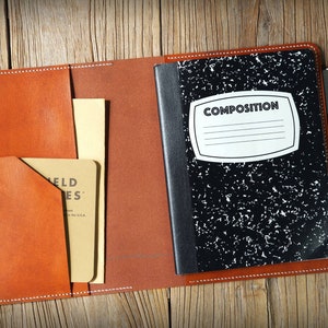Könnte beinhalten: Eine braune Leder-Notizbuchhülle mit einem schwarzen Kompositions-Notizbuch im Inneren. Die Hülle hat eine Tasche auf der Vorderseite und eine Klappe, die mit einem Druckknopf geschlossen wird. Auf der rechten Seite der Hülle befindet sich ein Stift im Stifthalter.