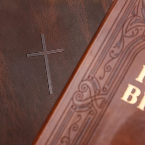 May include: Close-up of two brown leather-bound books. One features a debossed cross, the other has ornate gold lettering and decorative patterns. The books have a vintage, classic aesthetic.