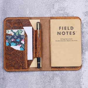 Può includere: Una copertina per quaderno in pelle marrone con un motivo floreale in rilievo. La copertina ha un portapenne, slot per carte e una tasca per un blocco note. La copertina è aperta per rivelare un quaderno del marchio Field Notes.