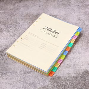 May include: A 2026 calendar planner refill with cream-colored pages featuring a personal information section. The planner has pre-punched holes on the left side and a set of colorful, protruding monthly index tabs labeled Jan through Dec.
