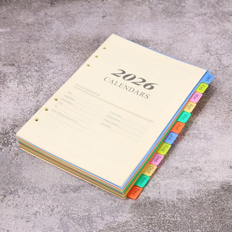 May include: A 2026 calendar planner refill with cream-colored pages featuring a personal information section. The planner has pre-punched holes on the left side and a set of colorful, protruding monthly index tabs labeled Jan through Dec.