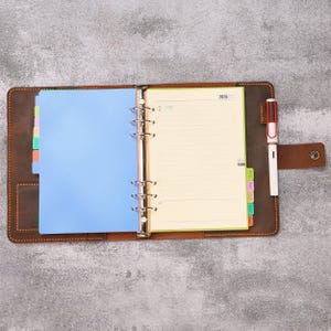 May include: A brown leather binder with a metal six-ring mechanism, containing a 2026 planner with cream-colored lined pages, a blue divider, and multi-colored index tabs. A white and brown pen is held in a side loop on the leather cover.