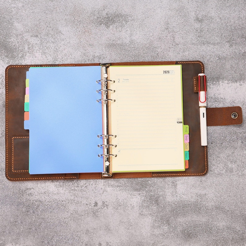 May include: A brown leather binder with a metal six-ring mechanism, containing a 2026 planner with cream-colored lined pages, a blue divider, and multi-colored index tabs. A white and brown pen is held in a side loop on the leather cover.