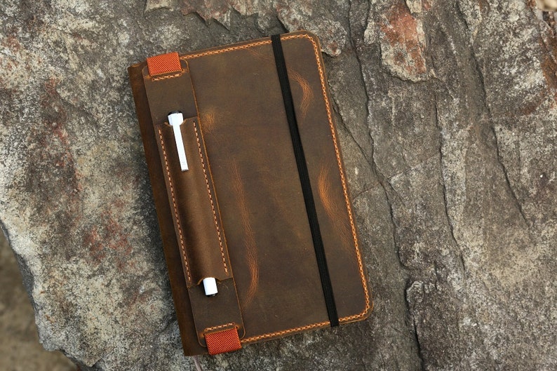 Puede incluir: Una cubierta de cuaderno de cuero marr&oacute;n con un portaplumas y un cierre el&aacute;stico. La cubierta tiene un aspecto desgastado y est&aacute; cosida con hilo naranja.