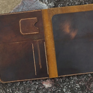 May include: A brown leather folder with a yellow leather interior. The folder has multiple pockets and a flap closure.