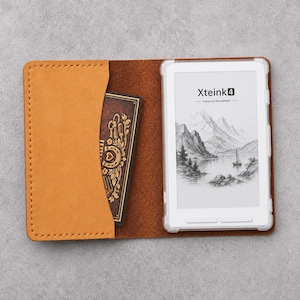 Può includere: Una custodia per e-reader in pelle color cuoio con una tasca laterale contenente una carta marrone scuro con illustrazioni dorate di fiori e ferri di cavallo. L'e-reader ha una cornice bianca e mostra il testo Xteink4 Focus on the present.