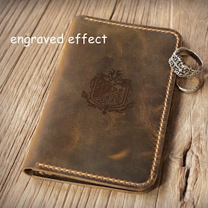 May include: A brown leather passport holder featuring a central engraved emblem and the text "engraved effect" in the upper left corner. The cover is stitched with light brown thread along the edges and is accompanied by two silver rings.