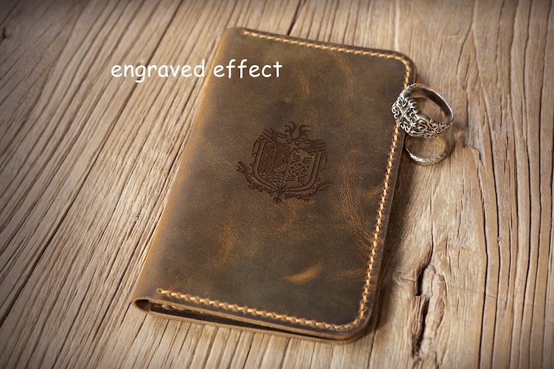 May include: A brown leather passport holder featuring a central engraved emblem and the text "engraved effect" in the upper left corner. The cover is stitched with light brown thread along the edges and is accompanied by two silver rings.