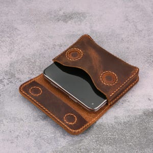 May include: A dark brown leather phone case with a flap closure featuring four circular stitched accents. The case is constructed with visible light-colored thread along the edges and holds a rectangular smartphone with a black screen.