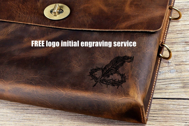 May include: A brown leather bag with a brass clasp and a decorative engraving of a feather and a crown of thorns. The text "FREE logo initial engraving service" is printed on the bag.