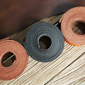 May include: Three rolls of leather in different colors: brown, black, and a lighter brown. The leather is rolled up and the edges are visible. The leather is on a wooden surface.