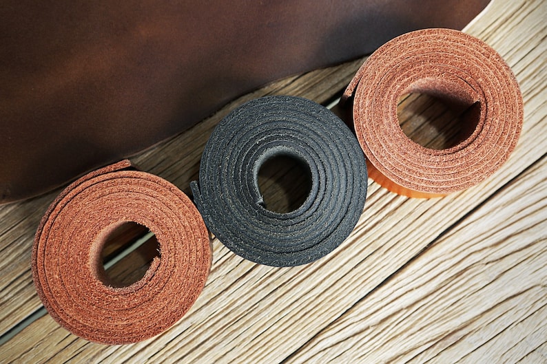 May include: Three rolls of leather in different colors: brown, black, and a lighter brown. The leather is rolled up and the edges are visible. The leather is on a wooden surface.