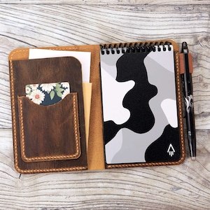 May include: A brown leather notebook cover with a spiral bound notebook inside. The notebook has a black and white camouflage pattern and a small arrow symbol. The cover has a pocket for cards and a pen holder.