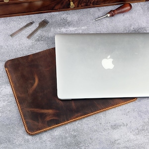 May include: A brown leather laptop sleeve with a laptop inside. The laptop is silver and has an Apple logo on the back.